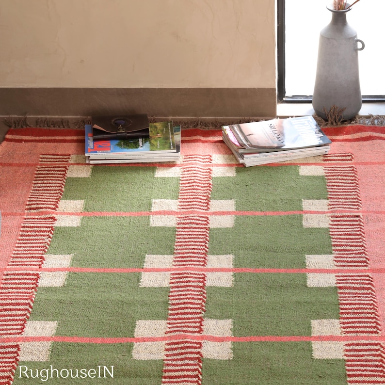 May include: A rectangular rug with a geometric pattern in shades of green, pink, and white. The rug features a central green field with white squares and pink horizontal stripes. Red and white striped borders frame the design. Magazines and a vase are on the rug.