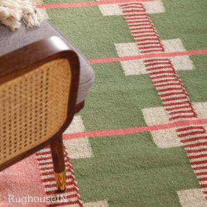 May include: A green and red patterned rug with a geometric design. The rug features a repeating pattern of red stripes and white squares on a green background. A wooden chair with a woven back is partially visible.