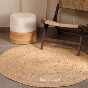 Pode incluir: Um tapete redondo de juta com um design espiral concêntrico está sobre um chão ladrilhado. Uma cadeira tecida com uma estrutura de madeira escura e um pufe cilíndrico bicolor, branco e bege, também estão na imagem. O tapete tem aproximadamente 120 cm de diâmetro.