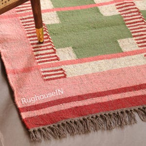 May include: A rectangular rug with a geometric design. The rug features a central green section with off-white and red accents. The border is pink and red with a fringe. The text "RughouseIN" is visible on the rug.