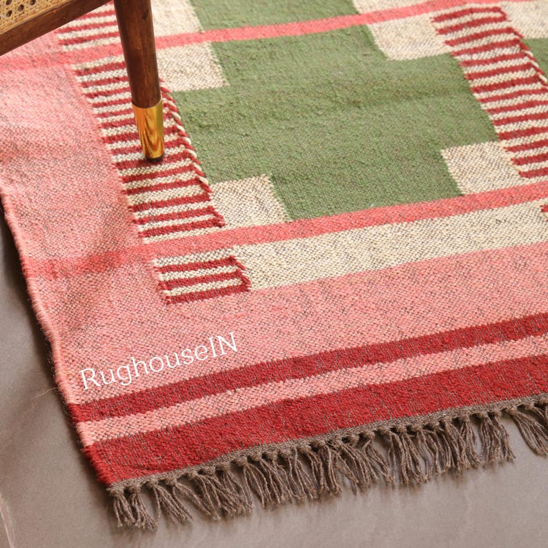 May include: A rectangular rug with a geometric design. The rug features a central green section with off-white and red accents. The border is pink and red with a fringe. The text "RughouseIN" is visible on the rug.