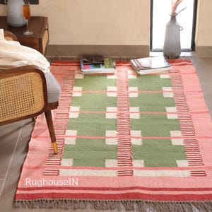 May include: A rectangular rug with a geometric pattern. The rug features a central green field with white squares and pink horizontal lines. The border is pink with red stripes and a fringed edge. The rug is placed on a floor near a chair.