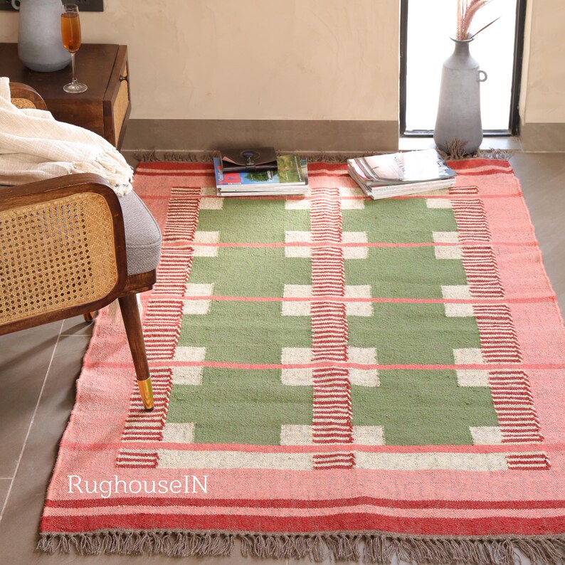 May include: A rectangular rug with a geometric pattern. The rug features a central green field with white squares and pink horizontal lines. The border is pink with red stripes and a fringed edge. The rug is placed on a floor near a chair.