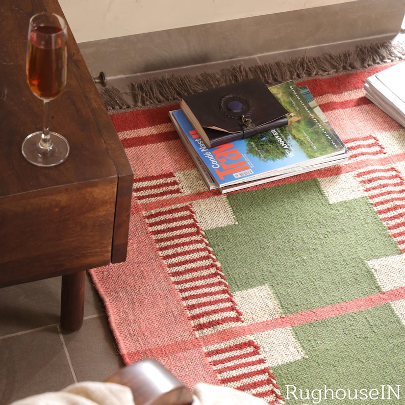 May include: A colorful geometric rug with a red, green, and white pattern. The rug is rectangular and has a fringe on one end. A dark wooden side table and a glass of wine are in the image.