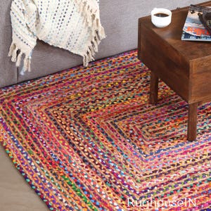 Può includere: Un tappeto in juta intrecciata multicolore con un motivo geometrico. Il tappeto presenta un vivace mix di tonalità rosse, blu, gialle e verdi, creando un design strutturato e accattivante. Il tappeto è posizionato su un pavimento accanto a un divano e un tavolino in legno.