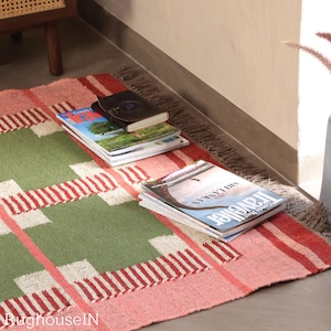 May include: A rectangular rug with a green and pink plaid pattern. Several magazines and a small book are stacked on the rug. The rug has a fringed edge and is placed on a concrete floor.
