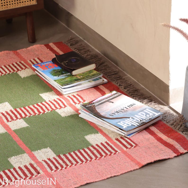May include: A rectangular rug with a green and pink plaid pattern. Several magazines and a small book are stacked on the rug. The rug has a fringed edge and is placed on a concrete floor.