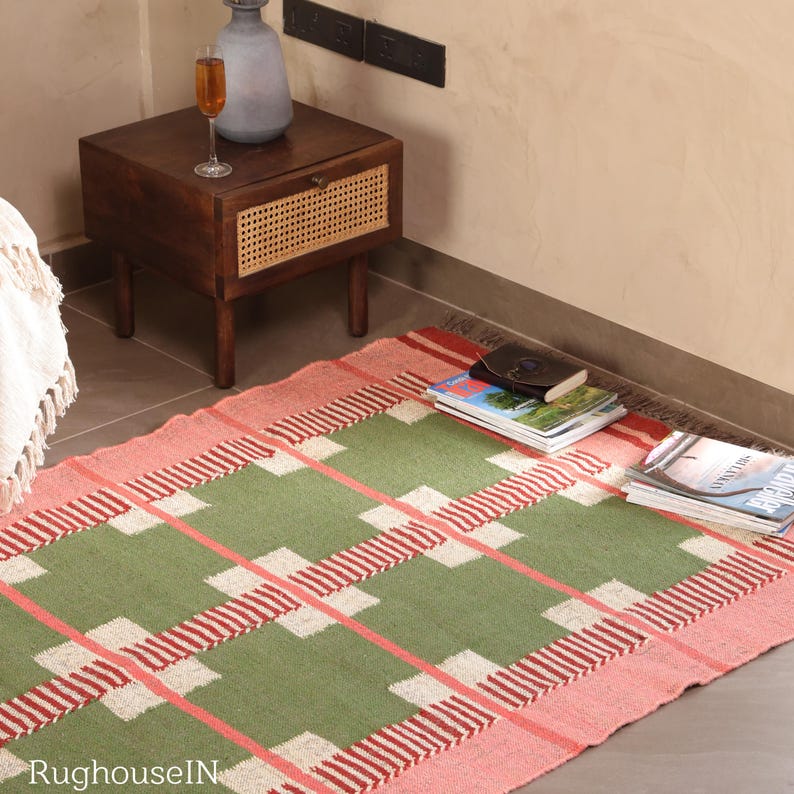 May include: A rectangular rug with a green and pink plaid pattern. The rug has a pink border and a central design of green squares and red stripes. The rug is placed on a tiled floor, with a wooden nightstand and books visible in the background.