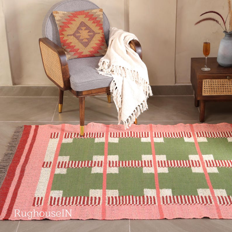May include: A pink and green rectangular rug with a geometric pattern is displayed on the floor. The rug has a fringed edge and is placed in front of a gray chair with a patterned pillow and a white throw blanket. The chair has wooden legs with gold tips.