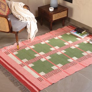 May include: A rectangular rug with a green and pink plaid pattern. The rug has a pink border with red stripes and fringe. The center of the rug is green with white and red accents. The rug is on a tiled floor.