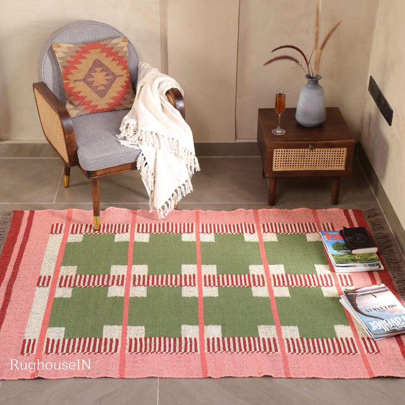 May include: A rectangular, hand-woven rug with a geometric pattern. The rug features a pink border, a green central field, and white and red accents. The rug is placed on a gray floor, with a chair, side table, and books nearby.