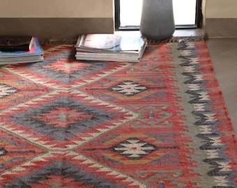 May include: A colourful rug with a geometric pattern in red, grey, and blue. The rug features diamond and zigzag designs, with the text "RughouseIN" in the bottom left corner. The rug is placed on a concrete floor.