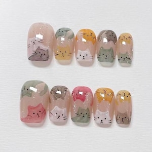 Puede incluir: Un conjunto de diez uñas artificiales, cada una pintada con un diseño diferente que presenta un gato de dibujos animados. Las uñas tienen forma ovalada y un acabado transparente y brillante. Los gatos son de varios colores.