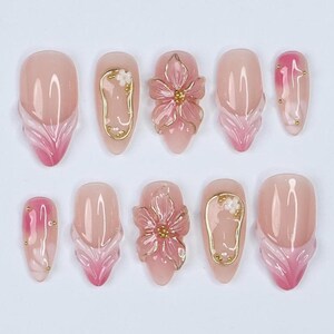 Puede incluir: Un conjunto de diez uñas artificiales con diversos diseños. Las uñas son principalmente rosas con detalles florales y abstractos. Algunas uñas presentan adornos florales en 3D, mientras que otras tienen detalles dorados y puntas rosas degradadas.