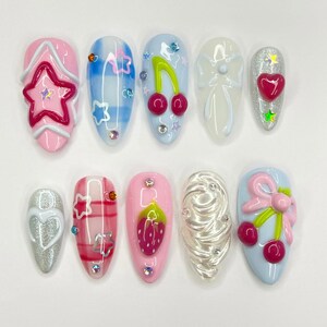 Puede incluir: Un conjunto de diez uñas artificiales con varios diseños. Las uñas presentan una gama de colores, incluyendo rosa, azul y plateado, y están adornadas con cerezas, estrellas, lazos y corazones. Algunas uñas tienen un acabado brillante, mientras que otras tienen adornos 3D.