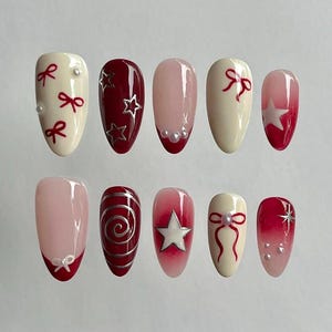 Puede incluir: Un conjunto de diez uñas artificiales con varios diseños. Las uñas presentan una mezcla de colores, incluyendo crema, rojo y rosa. Algunas uñas tienen detalles de lazo y estrella, mientras que otras tienen adornos de perlas y un diseño de remolino.