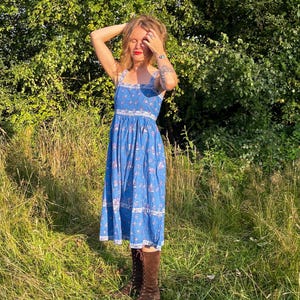 May include: A blue sundress with a floral pattern. The dress has a square neckline and a midi length. The person is wearing brown knee-high boots. The background is a field of tall grass and green trees.