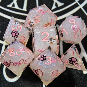 Puede incluir: Un conjunto de siete dados transparentes y brillantes con números rosas y diseños de huellas de patas. Los dados tienen varias formas, incluyendo un d20, d12, d10, d8, d6, d4 y un cubo. Para juegos de mesa.