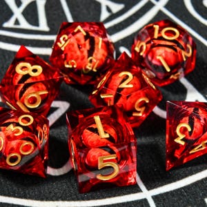 May include: A set of seven polyhedral dice with a red and black swirling pattern. The dice have gold-colored numbers and are designed for tabletop role-playing games. The dice include a D4, D6, D8, D10, D12, D20, and a percentile die.