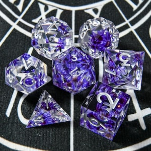 Puede incluir: Juego de siete dados de resina transparente con flores moradas incrustadas y numeración plateada. Los dados incluyen un d4, d6, d8, d10, d12, d20 y un dado de porcentaje. Los dados están colocados sobre una superficie con estampado en blanco y negro.