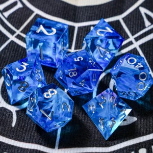 Op de afbeelding: Een set van zeven blauwe en zilverkleurige polyhedrale dobbelstenen. De dobbelstenen zijn transparant met een gemarmerd effect. Elke dobbelsteen heeft zilveren cijfers. De dobbelstenen liggen op een zwarte ondergrond met een wit geometrisch patroon.