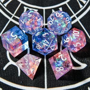 Puede incluir: Un conjunto de siete dados poliédricos en tonos azules, rosas y morados. Los dados tienen numeración blanca y un efecto brillante e iridiscente dentro de la resina transparente. Diseñados para juegos de mesa.