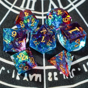 Puede incluir: Juego de siete dados poliédricos con una combinación de colores azul, morado y rojo. Los dados tienen números dorados y un efecto iridiscente brillante. Los dados están diseñados para juegos de rol de mesa.