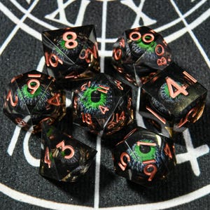Puede incluir: Juego de siete dados poliédricos con base negra y números color cobre. Cada dado presenta un diseño detallado de ojo verde y azul. Diseñado para juegos de rol de mesa.