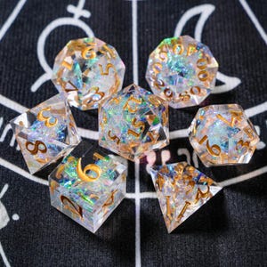 Könnte beinhalten: Ein Set aus sieben transparenten Harzwürfeln mit goldenen Zahlen und schillernden Glitzer-Einschlüssen. Die Würfel haben verschiedene polyedrische Formen, ideal für Tabletop-Spiele.