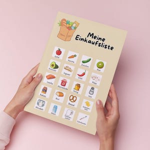 Może przedstawiać: Beżowa lista zakup&oacute;w zatytułowana "Meine Einkaufsliste" czarnym pismem, trzymana w dłoniach na r&oacute;żowym tle. Lista zawiera ilustracje artykuł&oacute;w spożywczych, takich jak jabłko, awokado i cukinia. Lista zawiera tekst "Apfel", "Karotte", "Zucchini", "Kiwi", "Avocado", "Ingwer", "Chili", "Pilze", "Lachs", "Marmelade", "Honig", "Salz", "Kakao", "Brot", "Brezel", "Scheiben-k&auml;se", "Windeln", "Milch", "Toilettenpapier" i "Zahnb&uuml;rste".