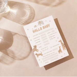 May include: A baby shower game card with the text "Hallo Baby" and illustrations of an elephant, giraffes, and leopards. The card is light beige with a brown backing and is on a light beige surface.