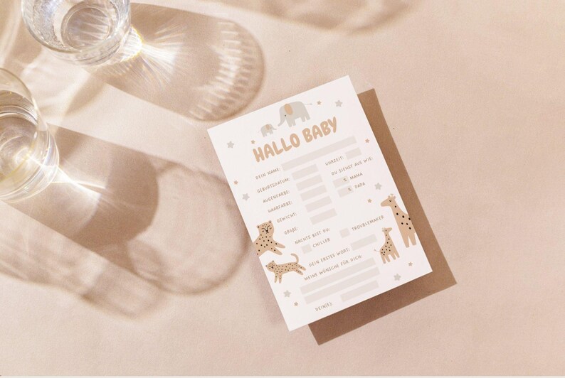 May include: A baby shower game card with the text "Hallo Baby" and illustrations of an elephant, giraffes, and leopards. The card is light beige with a brown backing and is on a light beige surface.