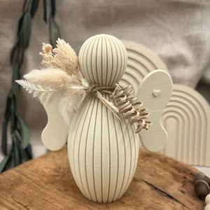 Può includere: Una statuina di angelo in legno color crema con corpo e ali a coste, adornata con fiori secchi e legata con spago. Un piccolo cuore è scolpito in un'ala. L'angelo è esposto su una superficie di legno.