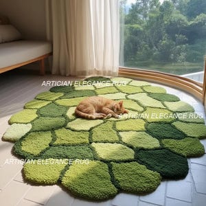 Könnte beinhalten: Ein runder, grüner Teppich mit einem einzigartigen, strukturierten Design. Der Teppich besteht aus verschiedenen Grüntönen und erzeugt ein mosaikartiges Muster. Eine rot getigerte Katze schläft auf dem Teppich. Der Text "ARTICIAN ELEGANCE RUG" ist auf dem Teppich aufgedruckt.