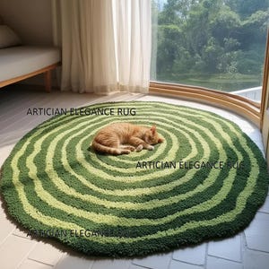 Könnte beinhalten: Ein runder, grüner Teppich mit konzentrischen Streifen in verschiedenen Grüntönen. Eine hellorange Katze schläft eingerollt in der Mitte des Teppichs. Der Text "ARTICIAN ELEGANCE RUG" ist auf dem Teppich aufgedruckt.