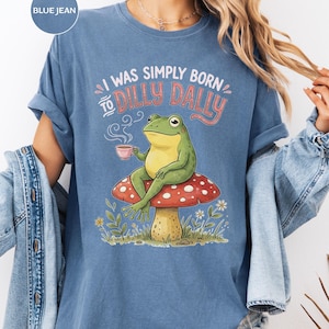 May include: A blue jean colored t-shirt featuring a graphic of a green frog sitting on a red mushroom with white spots. The frog holds a small pink cup with steam rising. The text reads I WAS SIMPLY BORN TO DILLY DALLY in pink and white letters.