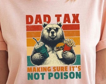 May include: A heather peach t-shirt featuring a graphic of a bear wearing sunglasses, holding a drink and a burger with fries. The text reads DAD TAX MAKING SURE IT'S NOT POISON, set against a background of horizontal stripes in red, orange, and yellow.