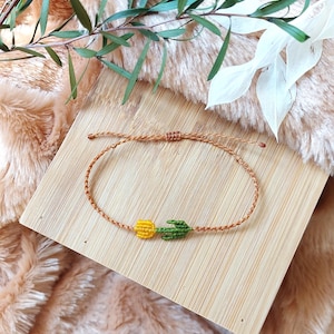 Pode incluir: Uma pulseira artesanal com um cordão castanho, uma flor amarela e um design de folha verde. A pulseira é apresentada numa superfície de madeira, com um fundo macio e texturizado e elementos decorativos.