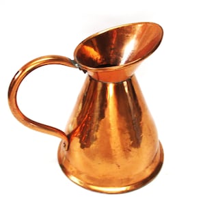 May include: A copper jug with a handle and a spout. The jug has a hammered texture and a shiny, reflective surface. The handle is curved, and the spout is wide and angled. The jug is likely used for pouring liquids.