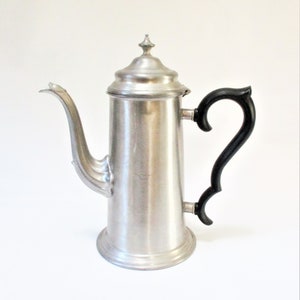 Könnte beinhalten: Eine hohe, silberfarbene Metall-Kaffeekanne mit gebogenem Ausguss und einem schwarzen, verzierten Griff. Die Kanne hat einen Deckel mit einem kleinen, dekorativen Knauf. Vintage und elegantes Design.