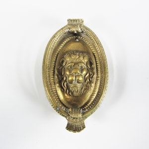Op de afbeelding: Een vintage messing ovale decoratie met een gedetailleerde leeuwenkop in het midden. Het gezicht van de leeuw wordt omgeven door een ovaal frame met een parelrand. Het heeft een kleine lus aan de bovenkant en een decoratieve basis.