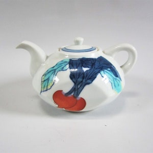Puede incluir: Tetera de cerámica blanca con tapa y asa. La tetera está decorada con un diseño de hojas azules y verdes y dos frutos rojos. La tetera tiene un pico y un asa curva.