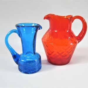 Puede incluir: Dos jarras de vidrio. Una es pequeña, azul vibrante con efecto craquelado. La otra es más grande, naranja brillante con un patrón de diamantes. Ambas tienen asas y picos, ideales para servir bebidas.