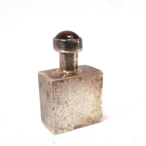 May include: A small, rectangular silver-toned bottle stopper with a dark, rounded top. The stopper has a simple, industrial design, with a cylindrical neck connecting the top to the rectangular base. The object is set against a white background.