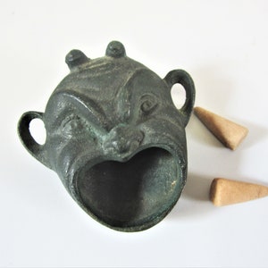 May include: A bronze-coloured incense burner shaped like a face with a wide open mouth and two small handles. Two brown incense cones are next to the burner. The burner has a distressed finish.
