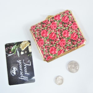 May include: A square, floral-patterned compact with a gold-colored frame, featuring red roses on a green background. A Kroger gift card with the text "Treat yourself" and two coins are also present.