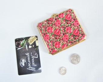 May include: A square, floral-patterned compact with a gold-colored frame, featuring red roses on a green background. A Kroger gift card with the text "Treat yourself" and two coins are also present.