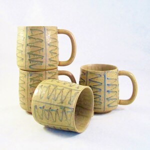 May include: Four beige ceramic mugs with a textured surface and a repeating blue and green zigzag pattern. Each mug has a rounded handle. One mug is lying on its side, while the others are stacked or upright.