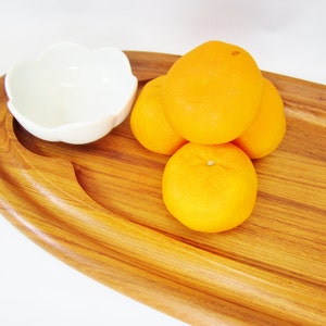 May include: A wooden serving tray with a white, flower-shaped bowl and several oranges. The tray has a natural wood grain finish. The oranges are bright orange and appear fresh.