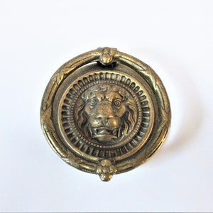 May include: Antique brass door knocker with a lion's head design. The round knocker features a detailed lion's face at the center, surrounded by a decorative ring with floral and leaf patterns. The knocker is a vintage piece, likely made of brass.